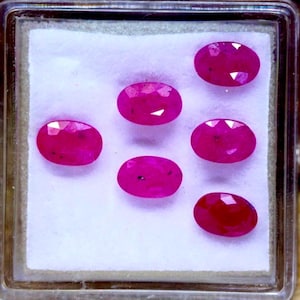 May include: Six oval-cut, vivid red gemstones are presented in a clear plastic case. The gemstones are set against a white background, highlighting their polished facets and rich colour. The gems are likely rubies.