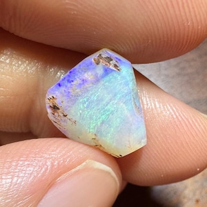 Könnte beinhalten: Ein kleiner, irisierender Opal mit einem weißen, blauen und grünen Farbmuster. Der Opal hat eine raue, natürliche Form und wird in einer Hand gehalten.