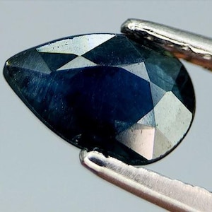 May include: A faceted, pear-shaped gemstone with a deep blue hue. The gem is held by a pair of silver tweezers, showcasing its polished surfaces and geometric cuts. The stone's facets reflect light, creating a sparkling effect.