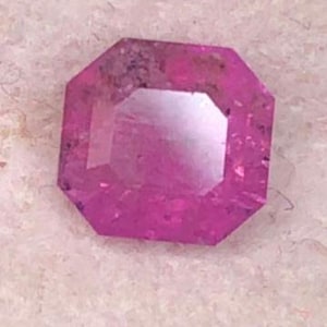 May include: A faceted, octagonal-cut, pink gemstone. The gem has a vibrant, saturated color and a reflective surface. The gemstone is set against a light-colored, textured background. The gem's facets catch the light, creating a sparkling effect.