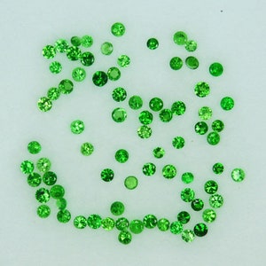 May include: A collection of small, round, green gemstones. The gemstones are scattered on a white surface.