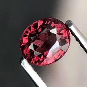 May include: A faceted, oval-shaped, deep red gemstone held by silver tweezers. The gem has multiple reflective surfaces, catching light and creating a sparkling effect. The background is a dark, blurred gradient.