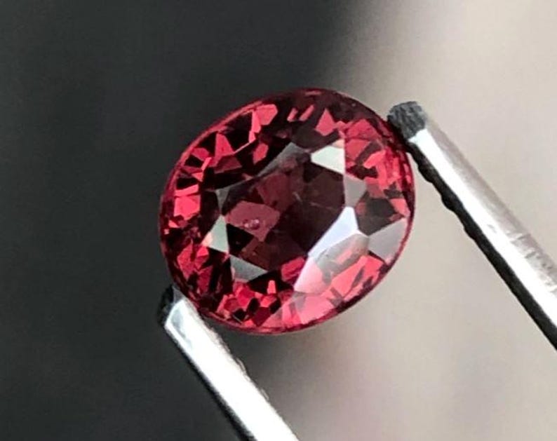 May include: A faceted, oval-shaped, deep red gemstone held by silver tweezers. The gem has multiple reflective surfaces, catching light and creating a sparkling effect. The background is a dark, blurred gradient.