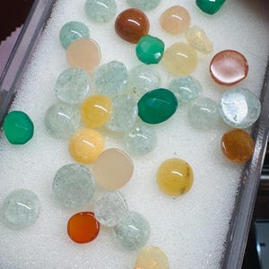 May include: Assortment of smooth, polished gemstones in various colors, including green, yellow, orange, and white. The stones are displayed in a white foam-lined tray.