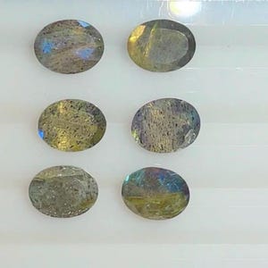 Könnte beinhalten: Sechs ovale Labradorit-Edelsteine. Die Steine zeigen eine Mischung aus Farben, darunter Blau, Grün und Gelb, mit einem schimmernden, irisierenden Effekt. Die Edelsteine ​​sind vor einem weißen Hintergrund angeordnet.