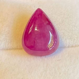 May include: A polished, teardrop-shaped gemstone in a vibrant magenta hue. The gem has a smooth, reflective surface, with subtle internal patterns. The stone is set against a soft, white background, highlighting its colour and form.