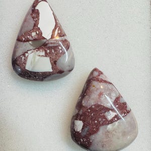 May include: Two teardrop-shaped gemstones in burgundy, white, and grey. The polished stones have a glossy finish. These gemstones are likely for jewellery or decorative use. Each stone is approximately 3 cm in length.