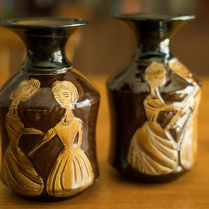 May include: Two brown ceramic vases with a dark glaze and gold painted designs of women. The vases have a wide mouth and a narrow neck.