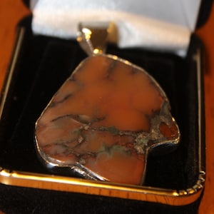 May include: A pendant with a large, irregularly shaped, orange-brown stone, set in a silver-colored metal frame. The stone has dark veins and is displayed in a black jewelry box with a white satin lining. The pendant is a unique piece of jewelry.
