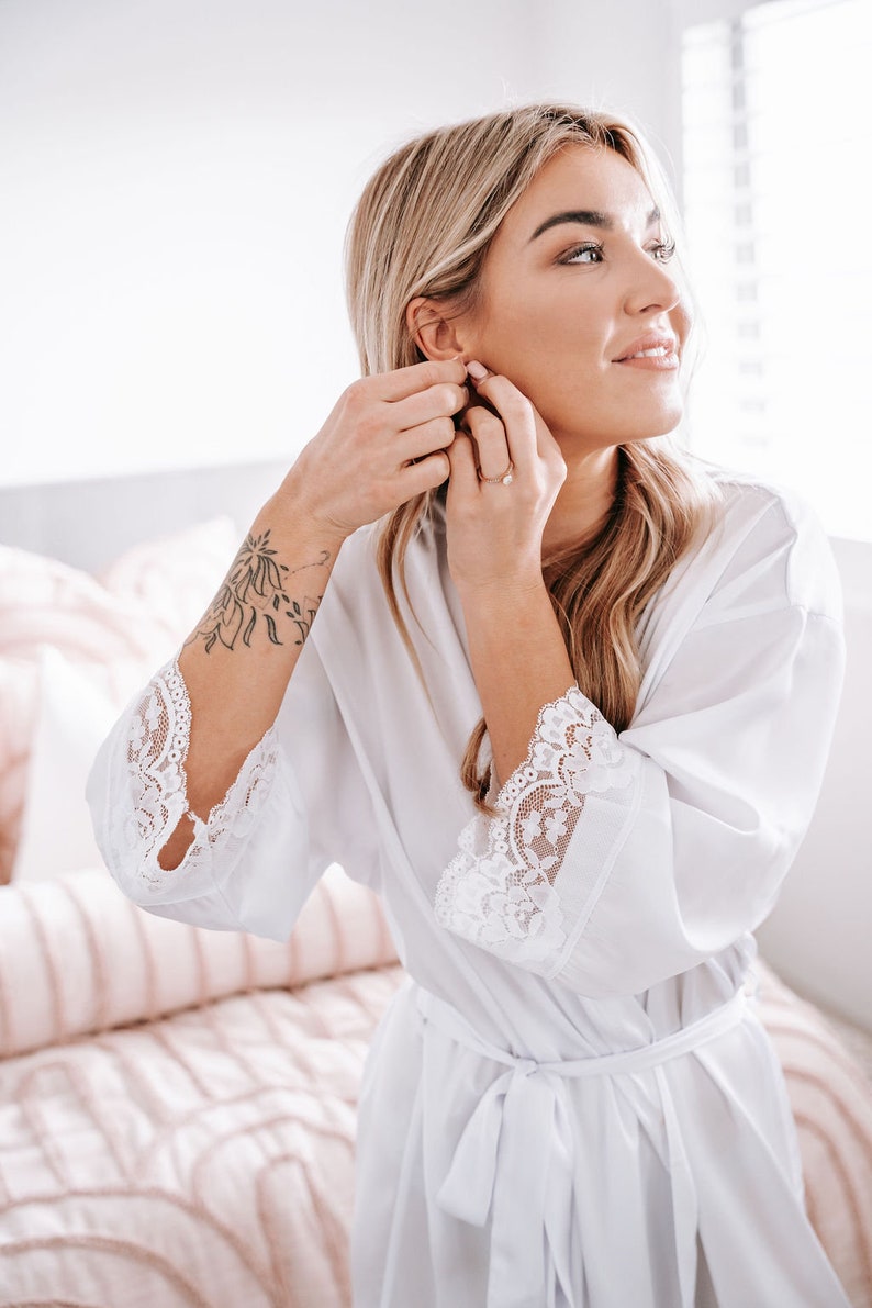 May include: A woman in a white satin robe with lace trim puts on earrings. The robe has a tie belt and the lace trim is on the sleeves and neckline.