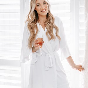 May include: A woman in a white robe with a ruffled hem holds a glass of champagne. She is standing in front of a window with white blinds.