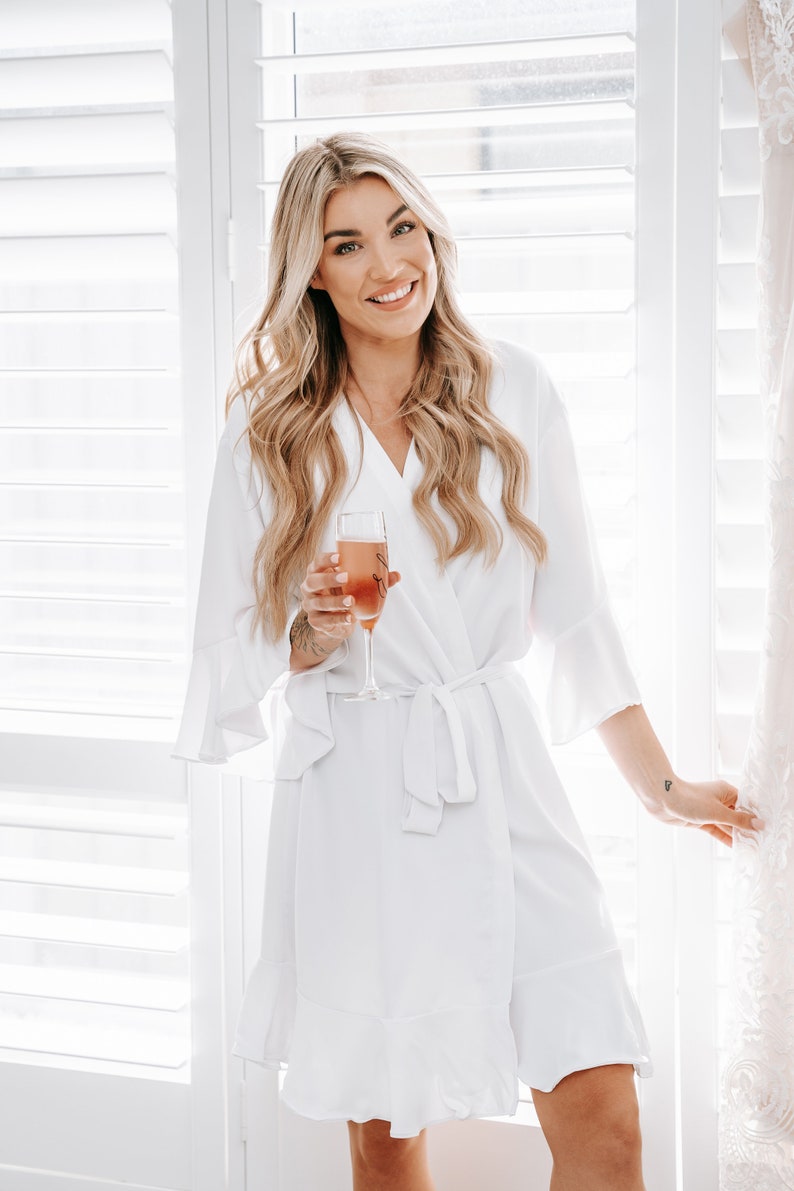 May include: A woman in a white robe with a ruffled hem holds a glass of champagne. She is standing in front of a window with white blinds.