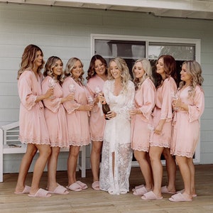 May include: A group of eight people wearing pink satin robes with lace trim and holding champagne flutes. The person in the center is wearing a white lace robe and holding a bottle of champagne.