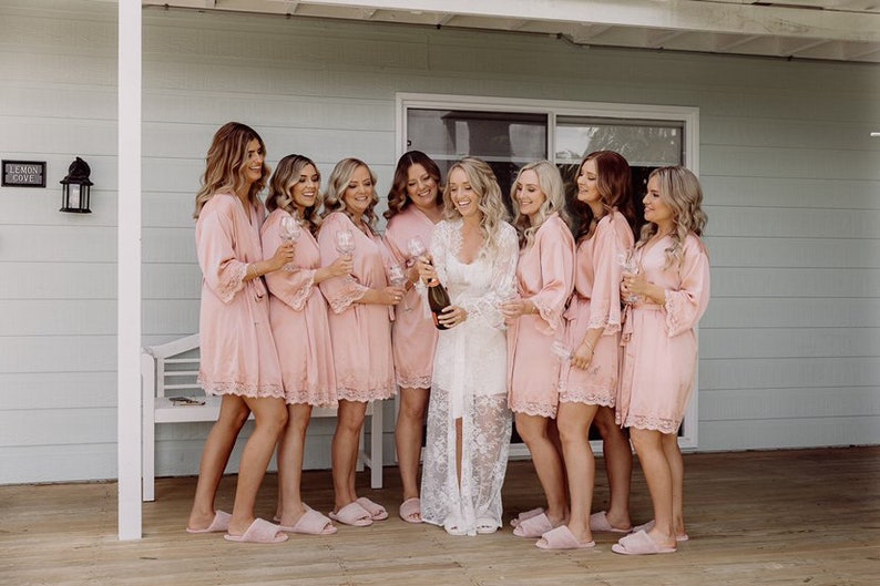 May include: A group of eight people wearing pink satin robes with lace trim and holding champagne flutes. The person in the center is wearing a white lace robe and holding a bottle of champagne.