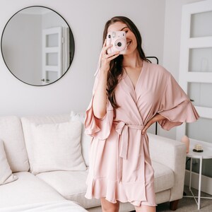 May include: A woman wearing a blush pink satin robe with a ruffled hem and a tie belt. She is holding a pink instant camera in front of her face.