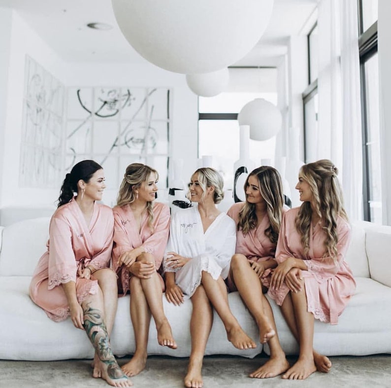 May include: Five women in pink satin robes with lace trim are sitting on a white couch. They are all smiling and looking at each other. The woman in the middle is wearing a white robe with lace trim. The women are in a bright, modern room with white walls and a large white balloon in the background.