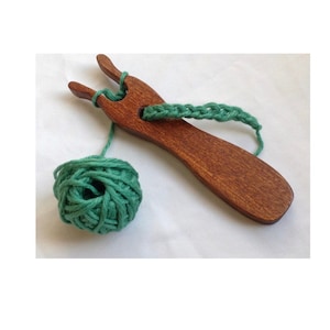 Puede incluir: Una horquilla de lucet de madera marrón oscuro con acabado liso, que presenta una parte superior de dos puntas y un orificio central. Una bola de hilo verde salvia está unida a la herramienta, con un tramo de hilo verde enhebrado por el orificio central.