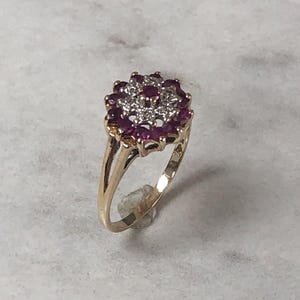 Op de afbeelding: Een gouden ring met een bloemmotief, met een cluster van kleine, ronde, dieprode edelstenen rond een centrale cluster van kleinere, heldere edelstenen. De ring heeft een delicate band.