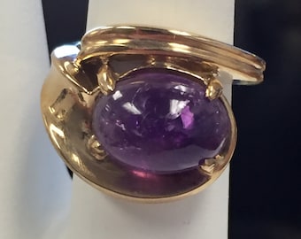 May include: A gold ring with a large, oval-shaped amethyst gemstone. The ring has a unique, curved design.