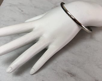 May include: A silver bangle bracelet with a minimalist design. The bracelet is a smooth, rounded band. The jewellery is displayed on a white mannequin hand against a marble-effect surface.