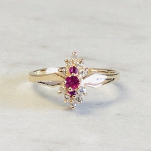 Könnte beinhalten: Ein goldener Ring mit einem Cluster aus kleinen weißen Steinen und einem größeren roten Stein in der Mitte.