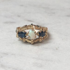May include: A gold ring featuring a white opal and two blue sapphires set in an ornate, decorative design.