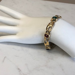 May include: A gold chain bracelet with three oval-shaped gemstones. The gemstones are red, green, and green.