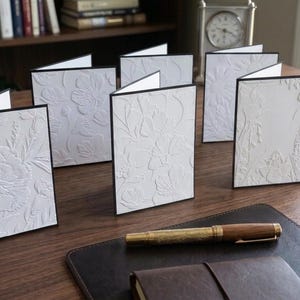 May include: A set of white folded greeting cards featuring embossed floral patterns on the front covers. Each card has a thin black border along the edges. The cards are arranged in a group on a wooden surface, accompanied by a wooden fountain pen.