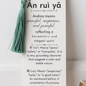May include: A rectangular white paper bookmark featuring black text that explains the meaning of the name Andrea in Chinese characters. The bookmark includes a sage green tassel attached through a hole at the top, with detailed definitions for the characters An, rui, and ya printed below the name.