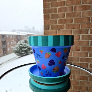 May include: A blue ceramic flower pot with a matching saucer, decorated with colorful illustrations of strawberries, blackberries, and blueberries. The rim features teal and green stripes. The pot is sitting on a mirrored surface.