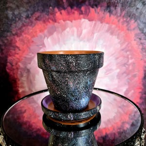 May include: A black and speckled ceramic flower pot with a matching saucer. The pot and saucer have a galaxy-like design with flecks of white, blue, and purple. The pot sits on a mirrored surface with a decorative black stand.