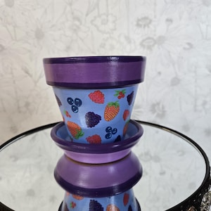 May include: A small, hand-painted ceramic flower pot and saucer set. The pot is light blue with a pattern of strawberries, blueberries, and blackberries. The rim and saucer are purple. The set is stacked on a decorative mirrored tray.