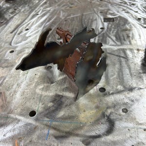 May include: A metal sculpture of the state of Michigan, with the Upper Peninsula and Lower Peninsula connected by a thin strip of metal. The sculpture is rusty and has a rough, industrial look.