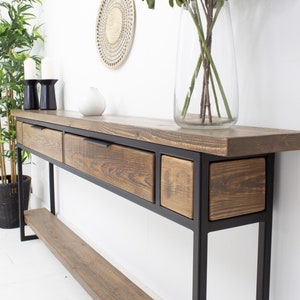 May include: A wooden console table with three drawers and a lower shelf. The table has a dark brown, textured top and drawer fronts, set within a black metal frame. A clear glass vase with flowers and decorative items sit on top.