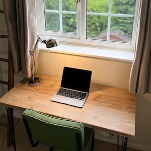 May include: A wooden desk with a laptop, a green chair, and a silver desk lamp. The desk is near a window with a view of greenery and is framed by beige curtains. The laptop is open, displaying a black screen.