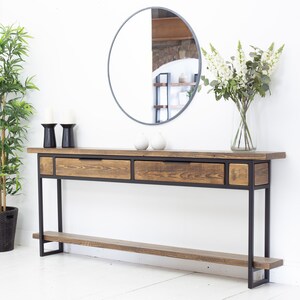May include: A wooden console table with a dark metal frame, two drawers, and a lower shelf. The table is paired with a round mirror, candles, and a vase of white flowers. The wood has a natural finish. The table's length is approximately 150 cm.