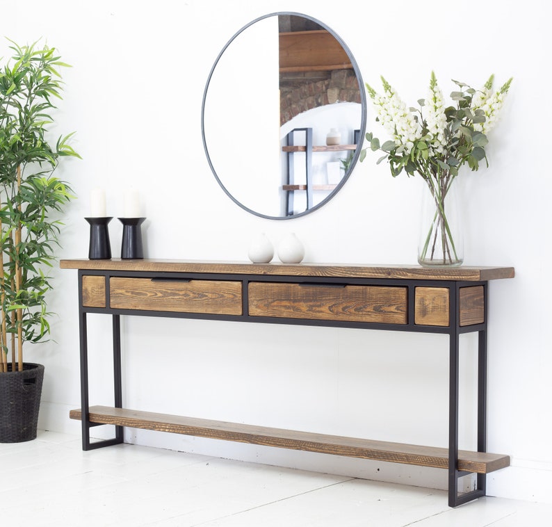 May include: A wooden console table with a dark metal frame, two drawers, and a lower shelf. The table is paired with a round mirror, candles, and a vase of white flowers. The wood has a natural finish. The table's length is approximately 150 cm.