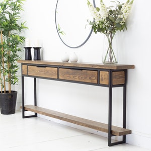 May include: A rustic console table with a dark stained wood top and drawers, supported by a black metal frame. The table features two drawers and a smaller compartment. A lower shelf runs the length of the table. The table is 152 cm long.