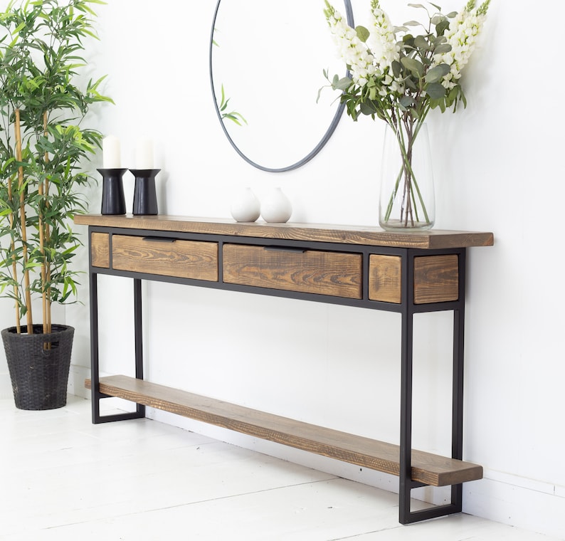 May include: A rustic console table with a dark stained wood top and drawers, supported by a black metal frame. The table features two drawers and a smaller compartment. A lower shelf runs the length of the table. The table is 152 cm long.