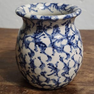 May include: A small, rounded ceramic vase featuring a cream-colored base with a mottled, sponge-painted pattern in deep blue. The vessel has a wide, flared rim and a bulbous body, showcasing a hand-applied finish with irregular blue markings.