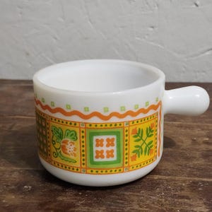 May include: A white milk glass mug with a single handle featuring a colorful repeating pattern of orange and green floral and geometric squares, accented by an orange wavy line border near the top rim of the cylindrical vessel.