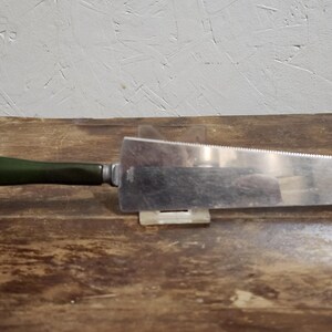 May include: A silver cake server with a green plastic handle. The server has a serrated edge and is resting on a wooden surface.