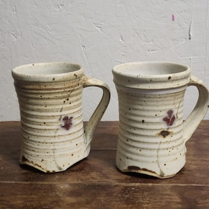 May include: Two ceramic mugs with a cream-colored glaze, featuring horizontal ribbed textures and brown speckles throughout. Each mug displays a small, hand-painted dark red floral motif on the front and includes a sturdy, curved handle.