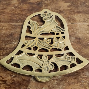 May include: A brass Christmas bell-shaped trivet with a holly and berry design. The trivet has a cutout pattern and a small loop at the bottom. The brass has a warm, golden hue, and the design is symmetrical.