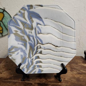 May include: An octagonal ceramic plate with a white base, featuring a blue and gray abstract design. The plate is displayed on a black stand, showcasing a unique artistic pattern. The plate's design resembles a landscape.