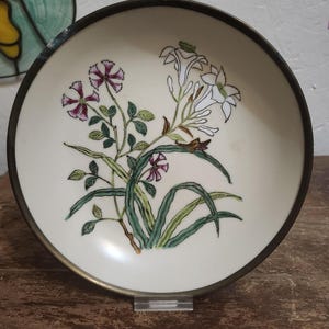 May include: A white ceramic bowl with a brass rim features a hand-painted floral design. The design includes white, pink, and green flowers with green leaves.