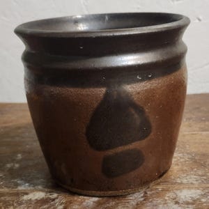 May include: A handmade, rustic ceramic pot with a dark brown rim and a lighter brown body. The pot features a dark brown, abstract design on the front. The pot is sitting on a wooden surface.