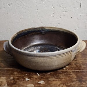 May include: A round ceramic bowl with two side handles, featuring a speckled beige exterior and a dark brown, glossy interior. The bottom of the bowl displays a subtle, embossed leaf pattern, and the rim is accented with a band of deep blue glaze.
