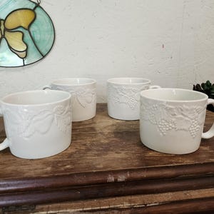 May include: Four white ceramic mugs with embossed grape and leaf designs are arranged on a wooden surface. Each mug has a curved handle. The mugs are of similar size and shape, suggesting they are part of a set.