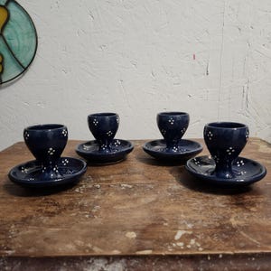 May include: Four navy blue ceramic egg cups with attached saucers. Each cup features a flared base and is decorated with small white polka dots. The egg cups are displayed on a wooden surface.